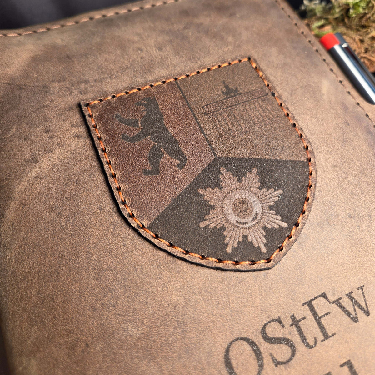 Handgefertigter DIN A5 Organizer aus braunem Leder mit aufgenähtem Wappen und feiner Naht in Orange