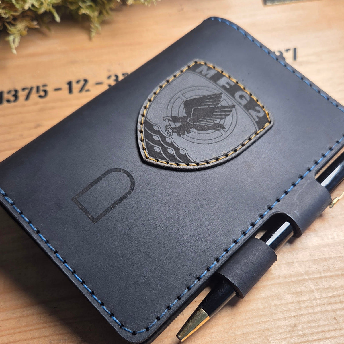 Handgefertigter Premium DIN A6 Organizer aus dunkelblauem Leder mit blauer Naht, aufgenähtem Wappen MFG 2 und integriertem Stiftehalter