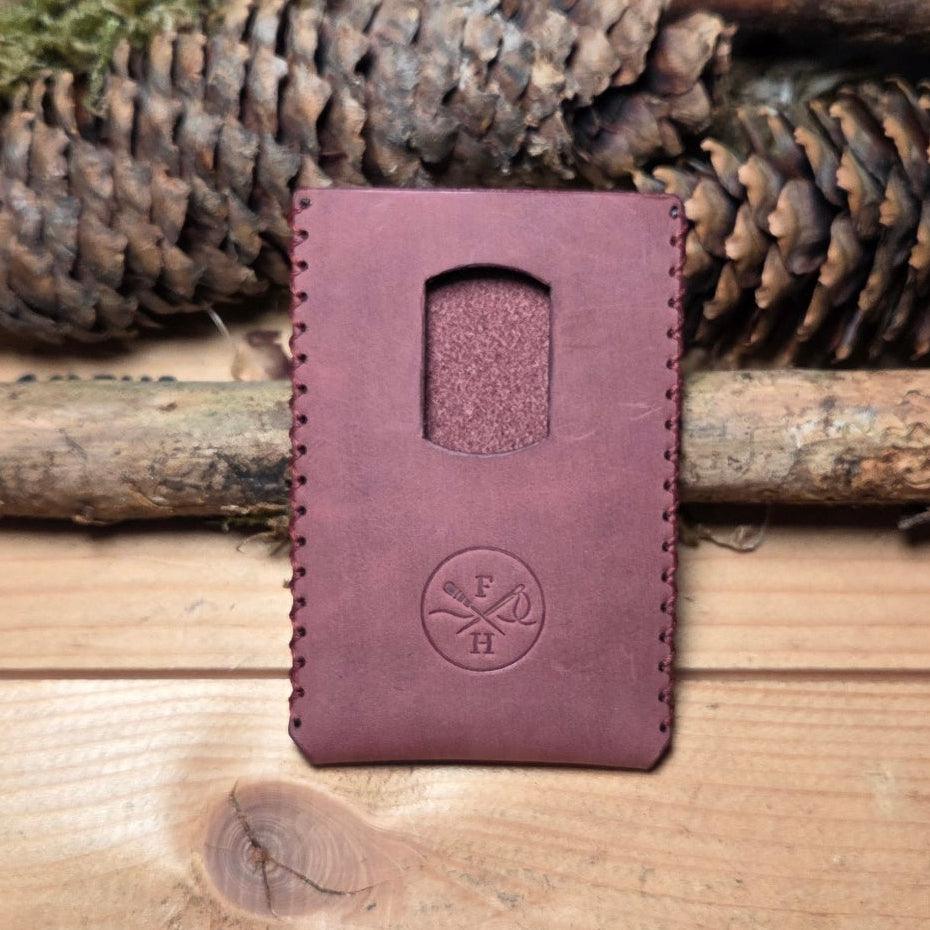 Slim Wallet aus Leder in dunklem Violett mit glatter Oberfläche, sichtbaren Handnähten und rundem Logo; oben ein Kartenauszug