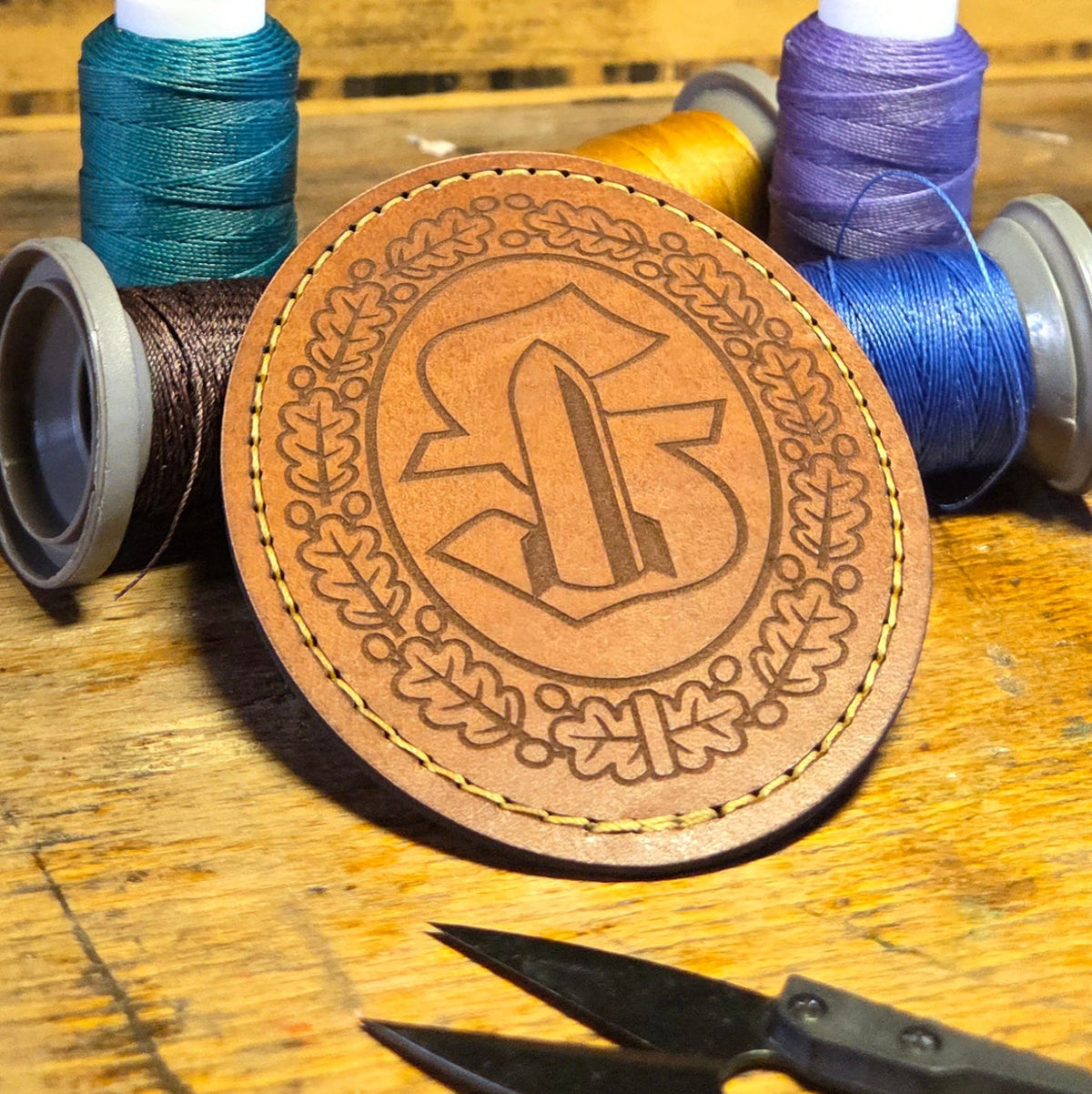 Runder Untersetzer aus Leder in braun mit gelber Naht und eingraviertem dekorativem Monogramm, umgeben von Eichenlaubmotiv